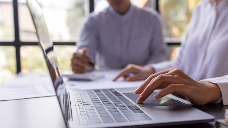 Pessoa usando notebook enquanto outra ao fundo consulta documentos, representando gestão e formalização de contratos intermitentes digitais em ambiente profissional.