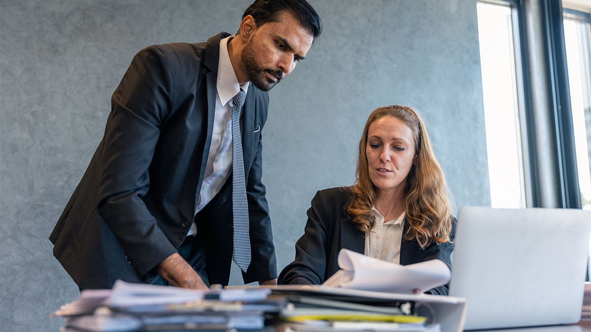 Duas pessoas vestidas com roupas formais em ambiente de escritório, uma mulher sentada diante de um laptop e papéis sobre a mesa, e um homem em pé ao lado dela. A cena sugere uma reunião ou análise de documentos, relacionada a processos trabalhistas.