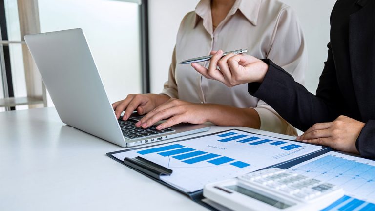 Duas pessoas sentadas à mesa de um escritório. Uma delas aponta para um gráfico de barras representando a transição da DIRF para o eSocial e os desafios de conformidade e segurança jurídica.