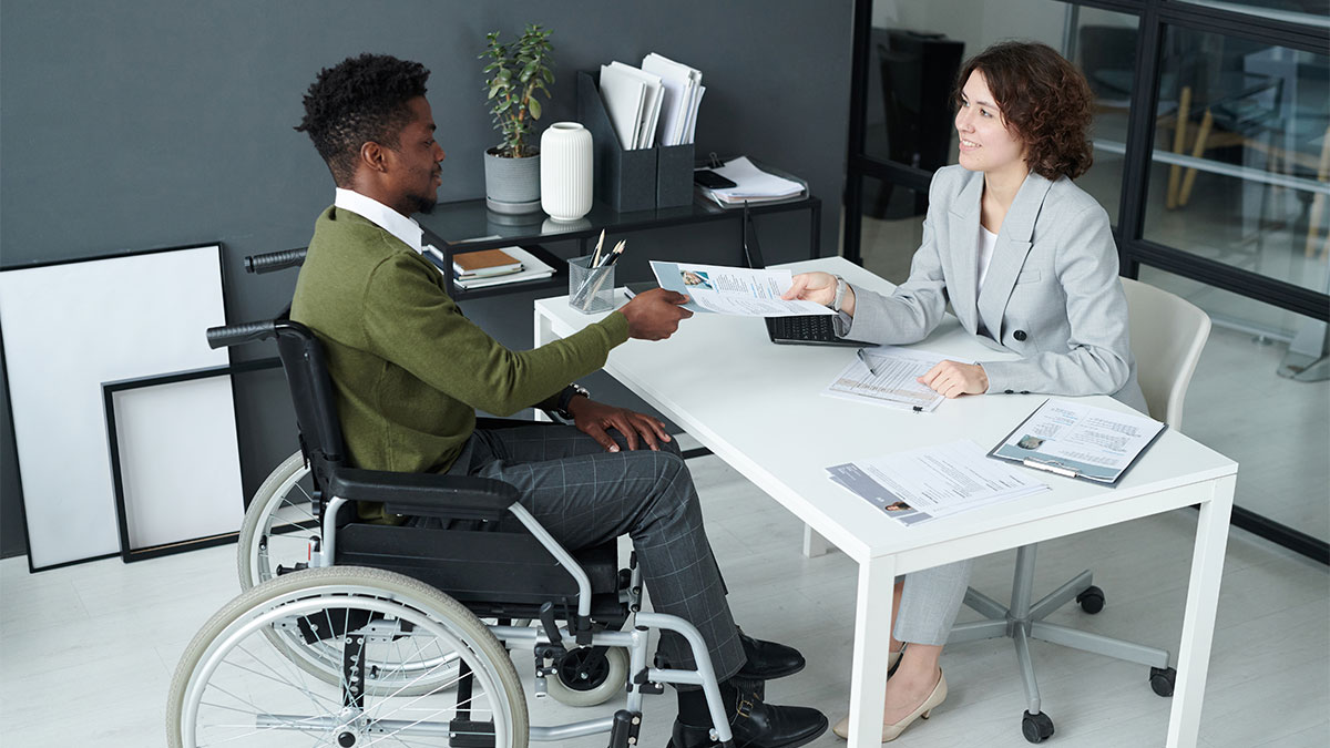 Pessoa em cadeira de rodas entregando documentos para advogada, representando a solicitação e atendimento do benefício BPC LOAS para pessoas com deficiência em situação de vulnerabilidade social.